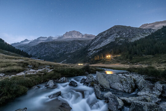 A scenic view of a mountain river flowing over rocks under a starry sky. Val di Fumo, Chiese river, Carr&egrave; Alto, Trentino. Lights from the Fumo Refuge are visible in the distance.