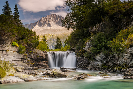 A scenic view of a waterfall cascading into a turquoise river, surrounded by lush greenery and mountains. Val di Fumo, Chiese river, Carr&egrave; Alto, Trentino. The water is smooth due to a long exposure.