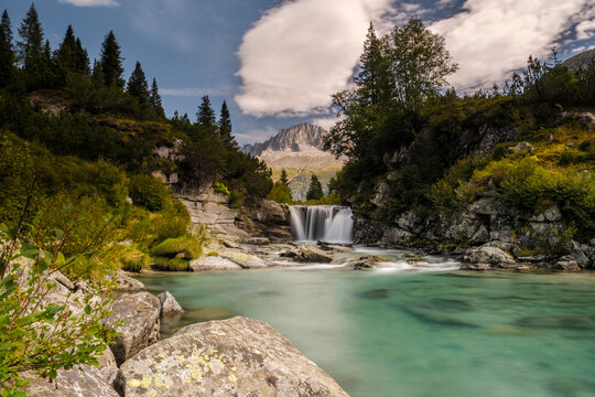 A scenic view of a waterfall cascading into a turquoise river, surrounded by lush greenery and mountains. Val di Fumo, Chiese river, Carr&egrave; Alto, Trentino. The water is smooth due to a long exposure.