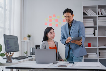 Obraz premium Two asian businesspeople are discussing and working together using laptop and tablet in modern office, analyzing financial statistics displayed on the computer screen