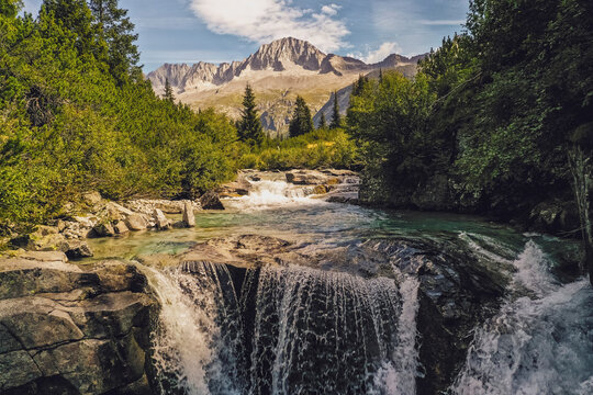 Val di Fumo, Chiese river, Carr&egrave; Alto, Trentino. A waterfall cascades over rocks into a clear pool, framed by lush greenery and a majestic mountain range in the background.