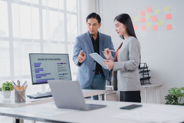 Two Asian businesspeople are discussing sales data displayed on a computer screen, collaborating on a project and analyzing market trends in a modern office environment