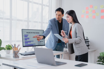 Naklejka premium Two asian businesspeople analyzing financial charts displayed on a computer screen, discussing strategies and collaborating on a project in a modern office environment