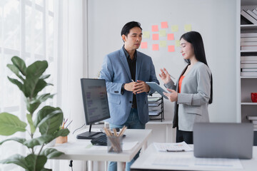 Two Asian businesspeople standing and discussing work while utilizing a tablet and sticky notes in a modern office, collaborating effectively on an important project