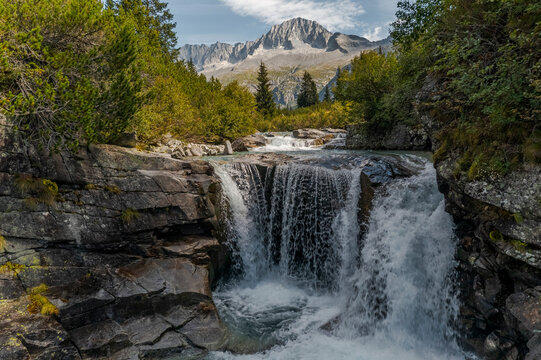 Waterfall cascading over rocks, surrounded by trees, with mountains in the background. Val di Fumo, Chiese river, Carr&egrave; Alto, Trentino. The water is clear and rushing, creating a scenic view.