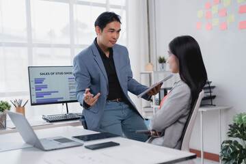 Fototapeta premium Two asian businesspeople are discussing sales data in the office, analyzing charts and reports on computer screen and tablet, collaborating on marketing strategy and planning