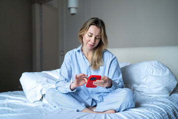 Serene middle aged woman in pajamas using smartphone while relaxing in bedroom. Interested female in sleepwear sits cross-legged on unmade bed checking phone after wake up. Morning with social media.