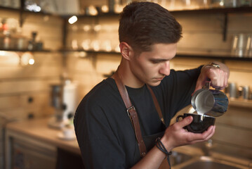 A young man with short hair pours steamed milk into a black cup. He is focused on creating latte art while working in a busy coffee shop filled with warm wooden decor and soft lighting