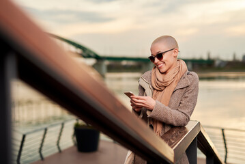 A woman with a bald haircut smiles while texting on her smartphone. She stands by a railing, wearing sunglasses and a warm scarf, with a beautiful waterfront view during sunset