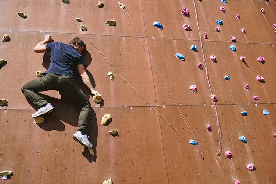 A man climbs an artificial rock wall with vibrant holds in an outdoor facility. He focuses intently while ascending under clear blue skies, embracing the joy of climbing