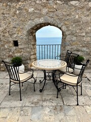 A photo showing a small round table and chairs with a small window through a fortress wall and a view of the Mediterranean Sea in Ulcinj, Montenegro.