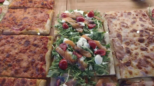 Close-up of assorted Italian pizza al taglio with plain cheese slices, tomato, arugula, mozzarella, and smoked salmon toppings in bakery display in Rome