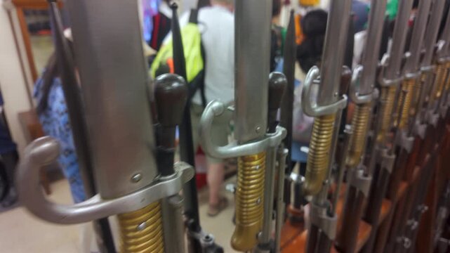 Close-up of vintage bayonets mounted on rifles in museum or military exhibition, with visitors blurred in background observing historical weapon collection