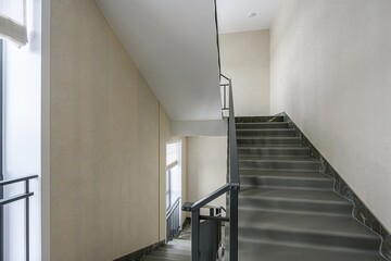 stairwell with grey stairs and black marble accents, leading upwards. Walls are light beige, and windows provide natural light