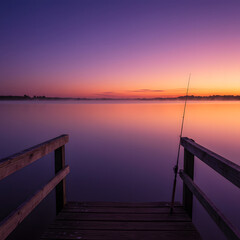 Fototapeta premium Serene Lake Sunset: A tranquil scene unfolds as dusk descends upon a calm lake, a fishing rod standing patiently on a wooden dock, creating a harmonious blend of nature and simplicity.