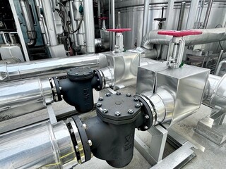 A close-up view of a complex network of industrial pipes, valves, and gauges. The large red handwheels and insulated metallic pipes highlight the intricate and heavy-duty nature of the cooling or heat