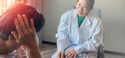 Asian female doctor in white coat consulting an elderly woman who is holding her head in pain, showing empathy and care in a bright clinic room, concept of healthcare, support, senior patient