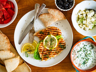 Greek style food: grilled chicken breast with lemon, pita bread,  tzatziki sauce on wooden table	