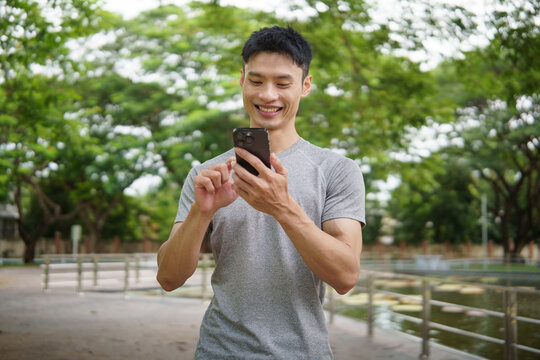 Smiling man in athletic wear holding smartphone while walking in a park. Healthy lifestyle concept. - Powered by Adobe