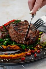 fried filet mignon steak on a plate on a pillow of greens with vegetables being cut with a knife