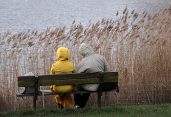 A couple sit on a bench by a lake in cold weather.