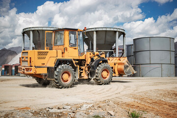 Heavy machinery operates at construction site with large loader moving earth near storage silos under clouds