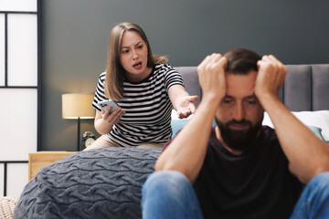 Fototapeta premium Betrayal. Angry woman with smartphone shouting at her offended husband on bed at home, selective focus