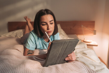A young woman is reading on her tablet while she is relaxing comfortably in bed