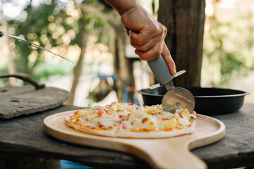 A hand using a pizza cutter to slice a pineapple pizza on a wooden cutting board outdoors.