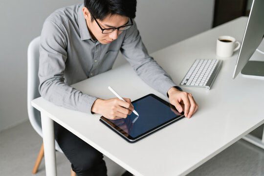 Young Asian man using digital tablet with stylus at modern office desk, working creatively and focused - Powered by Adobe