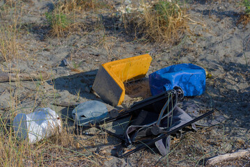 A pile of colorful plastic trash, including a yellow and blue container, is discarded on a sandy, overgrown beach.