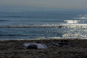 Two towels and a pair of shoes are seen left on a sandy beach next to the calm sea on a sunny day.