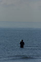 Fototapeta premium A lone man is seen standing in the sea, fishing with a long rod on a calm, misty day.