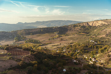 Countryside landscape with farmland, trees and mountains, with a cloudy blue sky in the background.