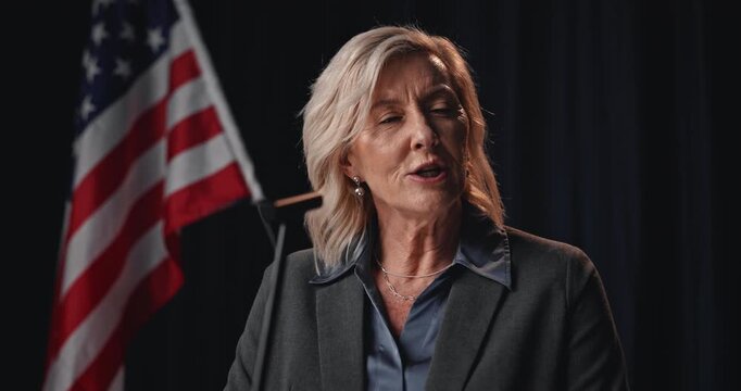 American flag, democracy and microphone with woman candidate at podium for political campaign. Election, government speech and politics with mature speaker in dark auditorium for democratic vote