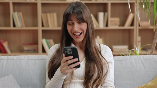Excited Caucasian woman raising fist in joy while holding smartphone and cheering with wide smile. Celebrating great news or reacting to message in cozy bright room with books and pillows around.
