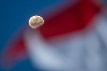 A photograph of a moon shining in the evening sky, with the waving flag of the Republic of Indonesia in the foreground, illustrates that the Republic of Indonesia is entering its independence month.