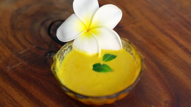 Closeup homemade ubtan facial in bowl with a big white champa flower. Ayurvedic face pack of Haldi (Turmeric), Rose water and gram flour. Natural beauty and skincare using since ancient India