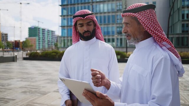 Two emirati businessmen wearing traditional saudi arabia clothing are posing together in front of modern glass buildings, creating a striking juxtaposition of tradition and modernity