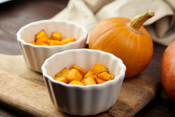 Freshly cut pumpkin cubes in white bowls on wooden cutting board, surrounded by whole pumpkins and soft fabric, showcasing seasonal ingredients for cooking and baking
