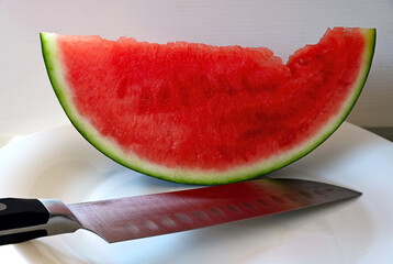 ripe, juicy red seedless watermelon slice macro on shiny white plate. large shiny steel vegetable knife in the foreground. thing green peel. healthy eating concept. vitamins and healthy eating.  