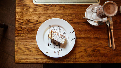 Delicious slice of cake topped with powdered sugar and chocolate sauce on a white plate,...