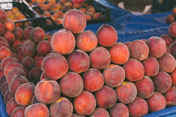 Fresh, organic peaches stacked in a market display. The peaches are ripe, with a vibrant orange-red color and a velvety texture. The setting is a local farmers' market.