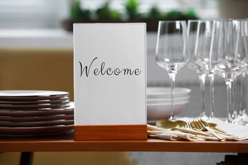 Holder with word Welcome, clean dishware and cutlery on wooden table in restaurant