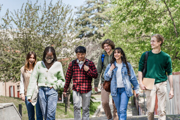 Happy diverse group of college students walking and laughing together on sunny campus path outdoors.