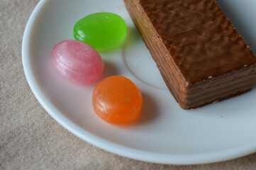 glass cup of black tea with a chocolate wafer and colorful hard candies on a white plate placed on soft beige fabric tea time aesthetics, cozy moments, dessert and confectionery, relaxation fall