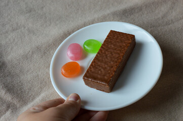 glass cup of black tea with a chocolate wafer and colorful hard candies on a white plate placed on soft beige fabric tea time aesthetics, cozy moments, dessert and confectionery, relaxation fall