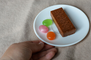 glass cup of black tea with a chocolate wafer and colorful hard candies on a white plate placed on soft beige fabric tea time aesthetics, cozy moments, dessert and confectionery, relaxation fall