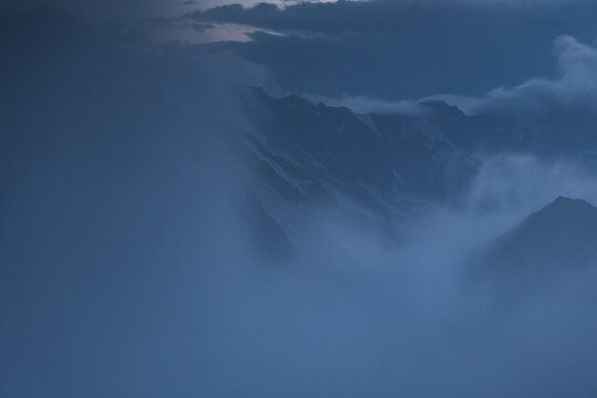 Misty mountain range in shades of blue. Clouds obscure the peaks, creating a sense of depth and mystery. Mantova refuge al Vioz, Peio, Trentino. 