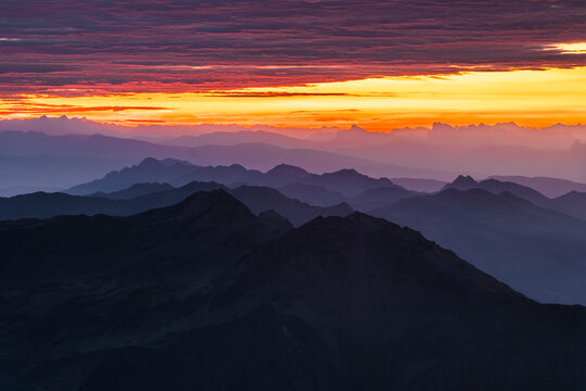 Layered mountain ranges fade into the distance under a vibrant orange and red sunset. Mantova refuge al Vioz, Peio, Trentino. The sky is streaked with colorful clouds.